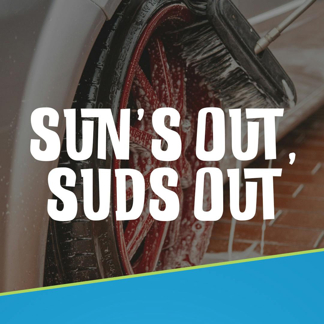 Text "Sun's Out, Sud's Out" in front of a black overlay and driver side wheel cover in suds being cleaned by a brush in the background