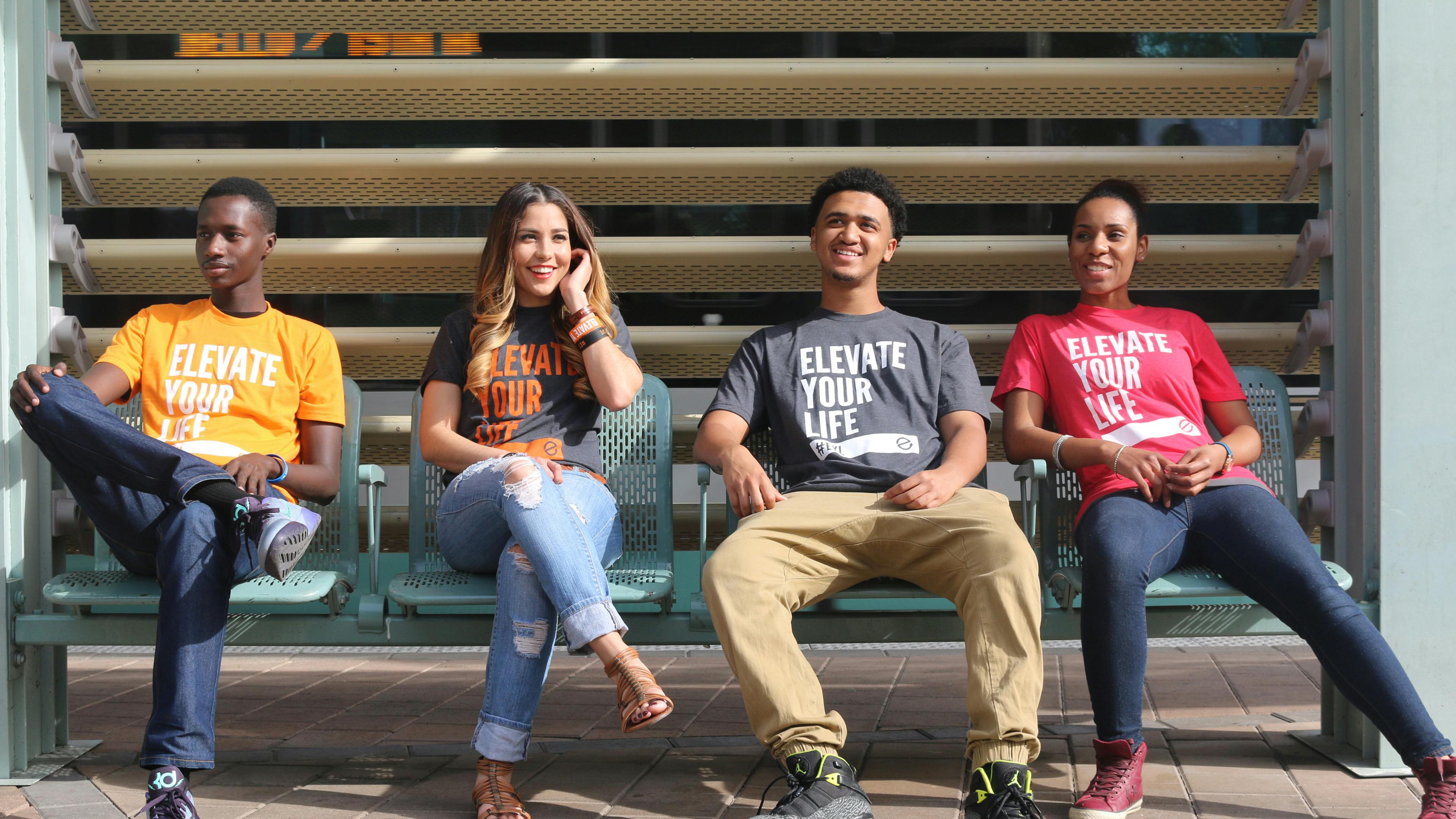 A group of people, 2 guys and 2 girls, sitting on a bus bench wearing different color Elevate Your Life shirts, all looking in different directions