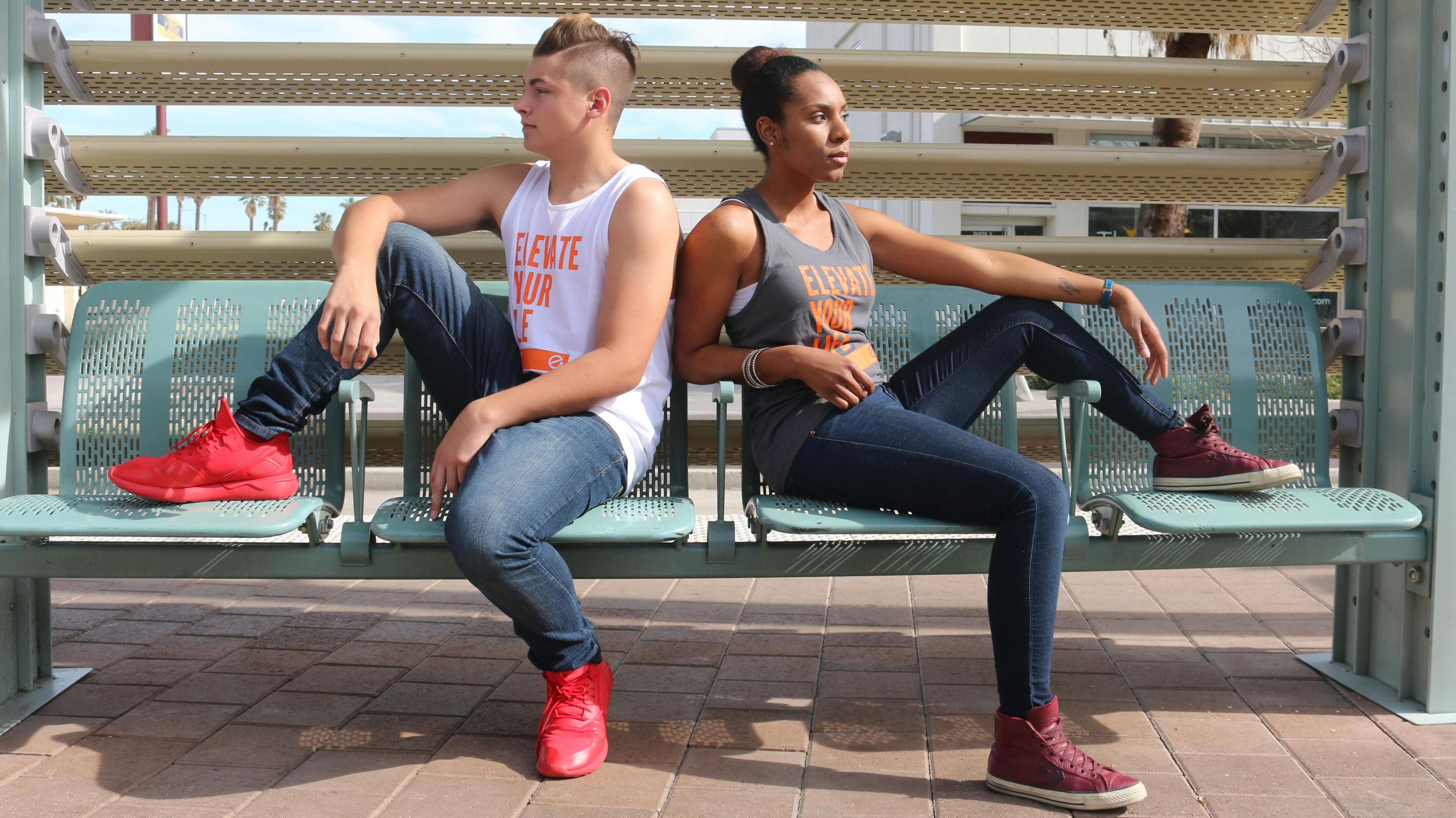 A guy and a girl back to back sitting on a bus bench wearing different color Elevate Your Life tanks
