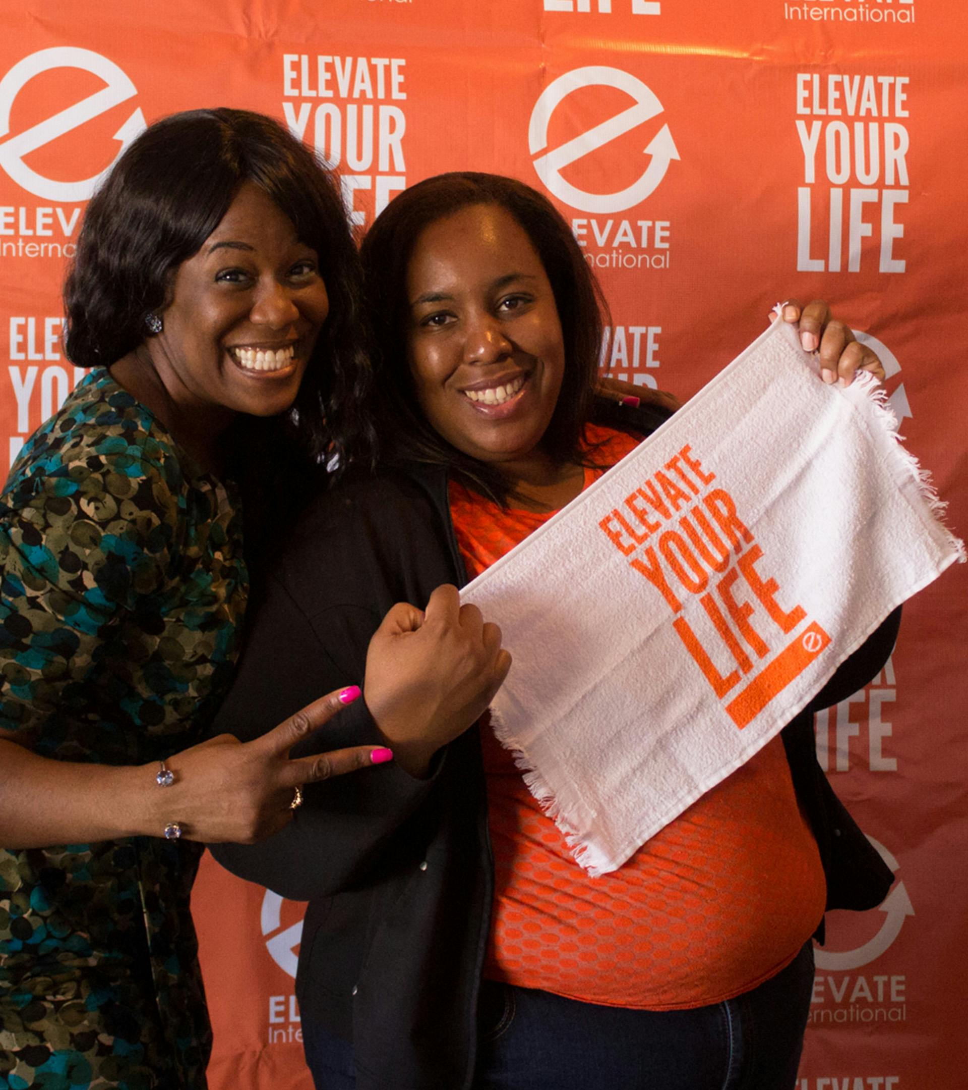 2 women smiling in front of a Step and Repeat for EI with one women holding a Elevate Your Life towel