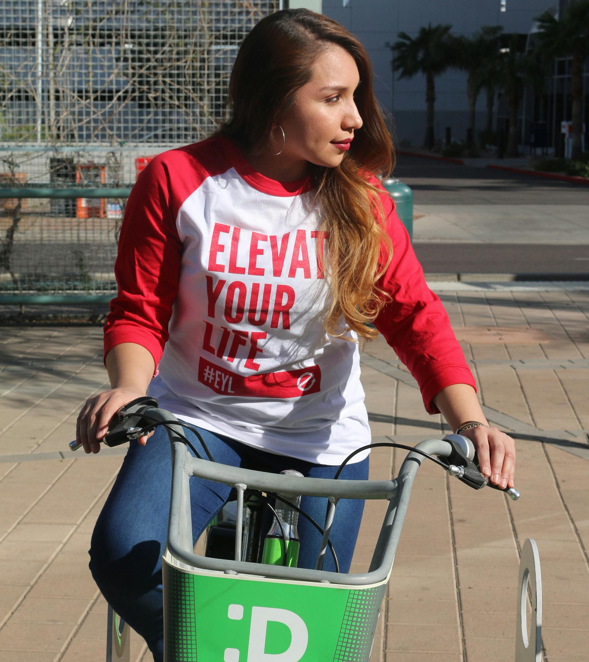 A woman on a bike wearing a red Elevate Your Life baseball tee looking to the left