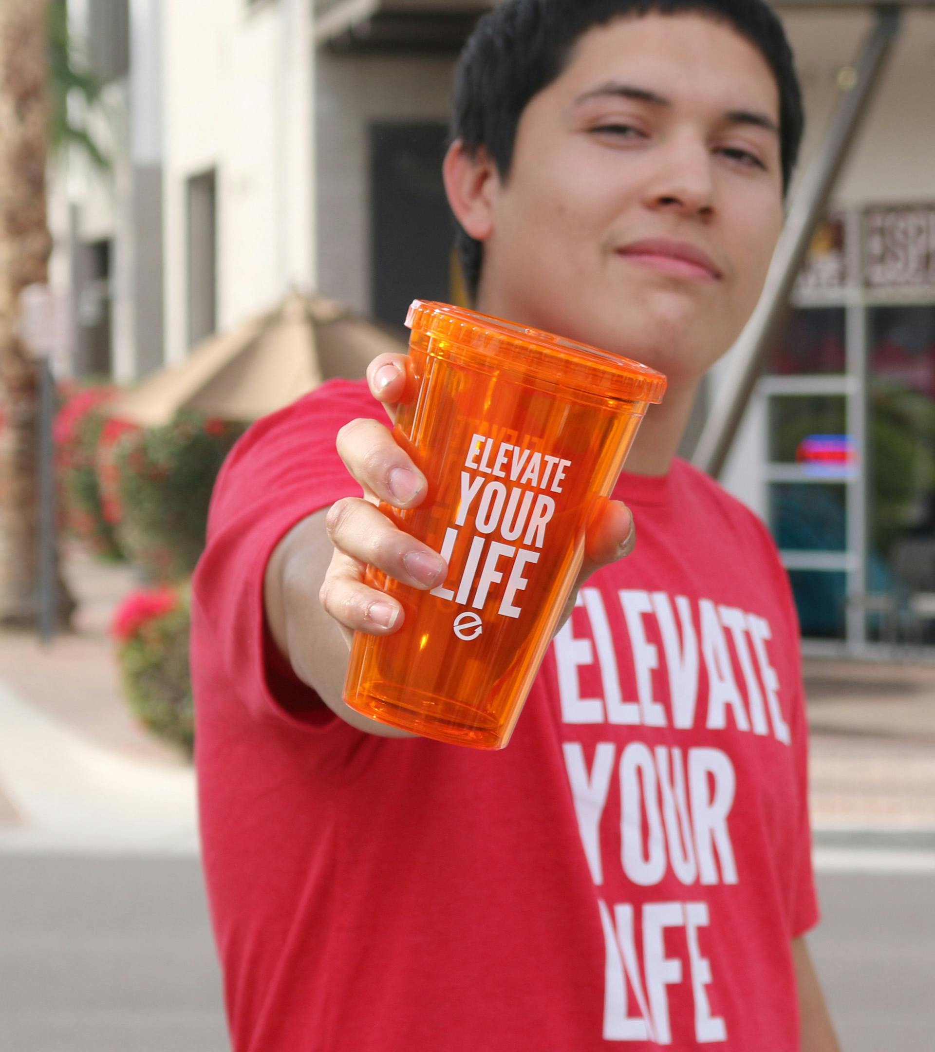 A guy who is slightly blurred holding an orange Elevate Your Life  tumbler in front that is in focus