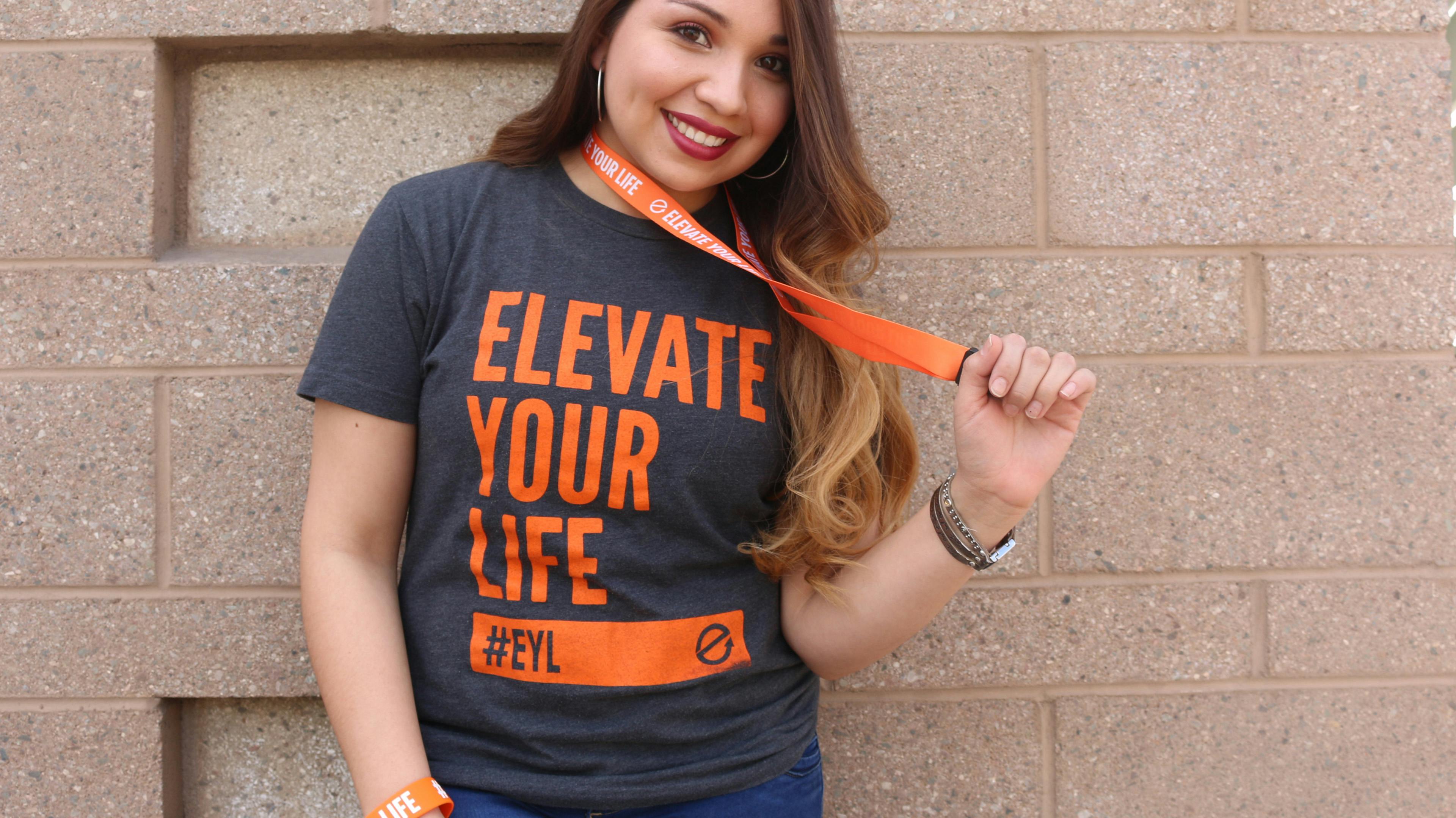 A woman wearing a charcoal gray and orange Elevate Your Life  shirt wearing an Elevate Your Life wristband and wearing a Elevate Your Life on her neck holding the end of the lanyard with her hand