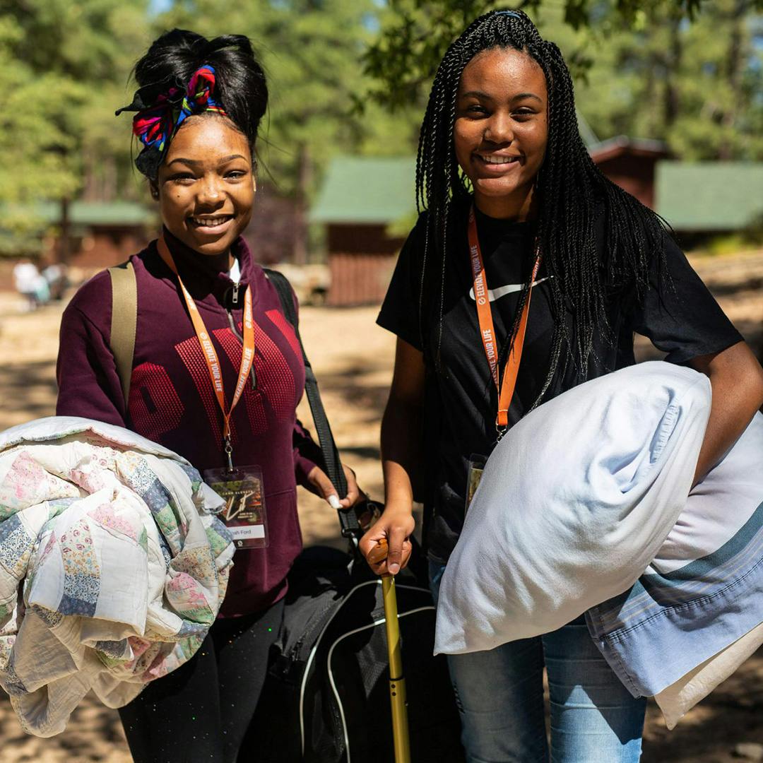 2 teenage women at camp smiling while carrying their luggage and pillows