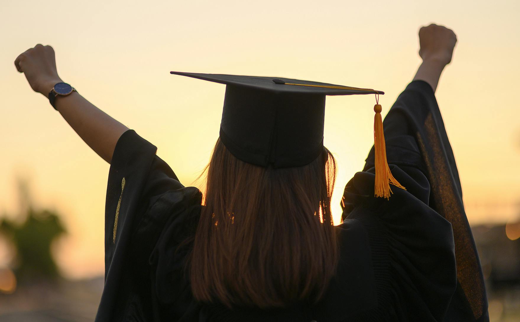 A girl facing away wearing her graduation hat and outfit with her arms raised in accomplishment